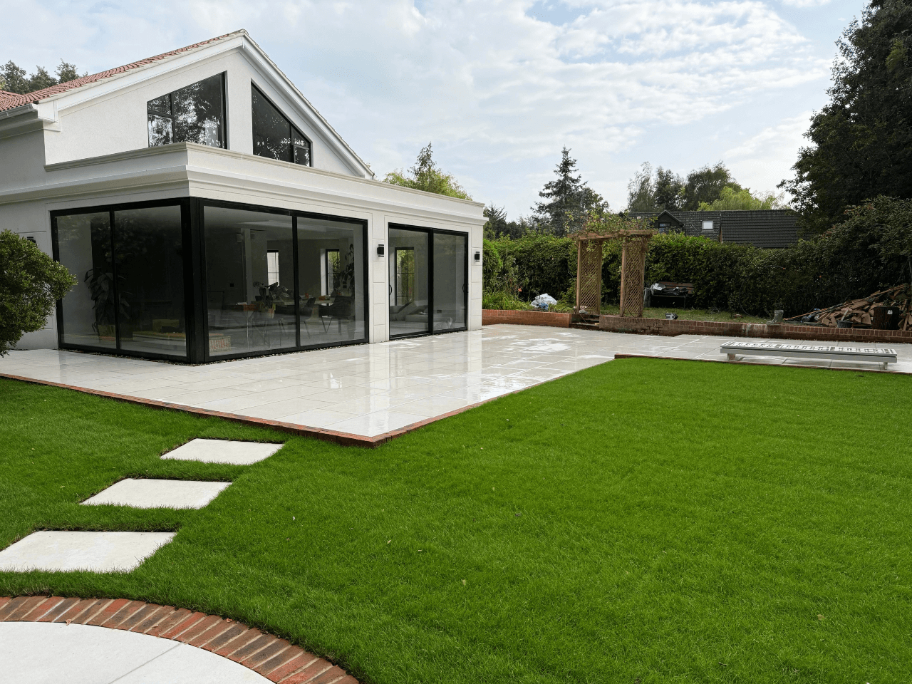 Modern white house with large black-framed sliding glass doors opening onto a tiled patio, overlooking a neatly landscaped garden with green lawn, stepping stones, and surrounding trees under a partly cloudy sky